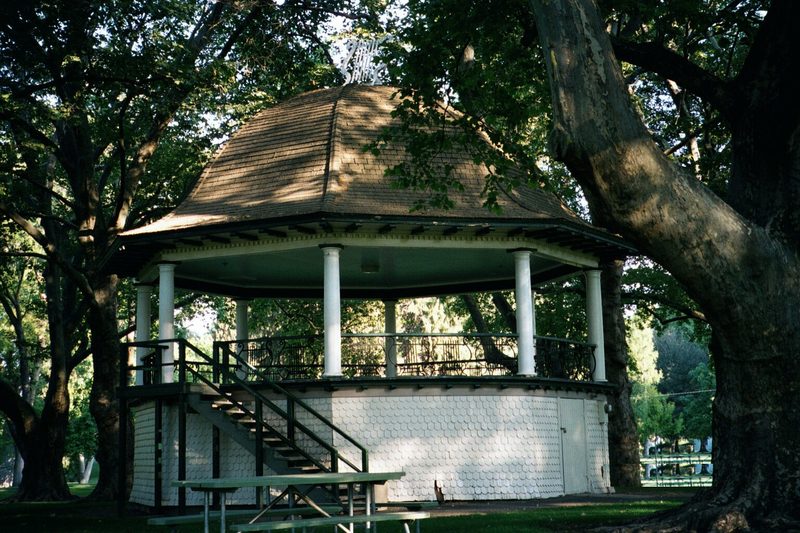 A white octagonal bandstand with a copper roof under large old trees in a park.
